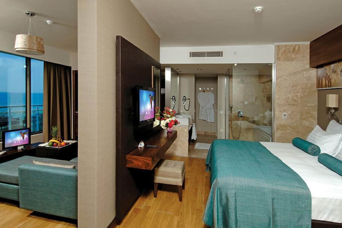 A modern hotel room with a large bed, a seating area with a TV, a shower area, and a balcony with a view of the ocean.