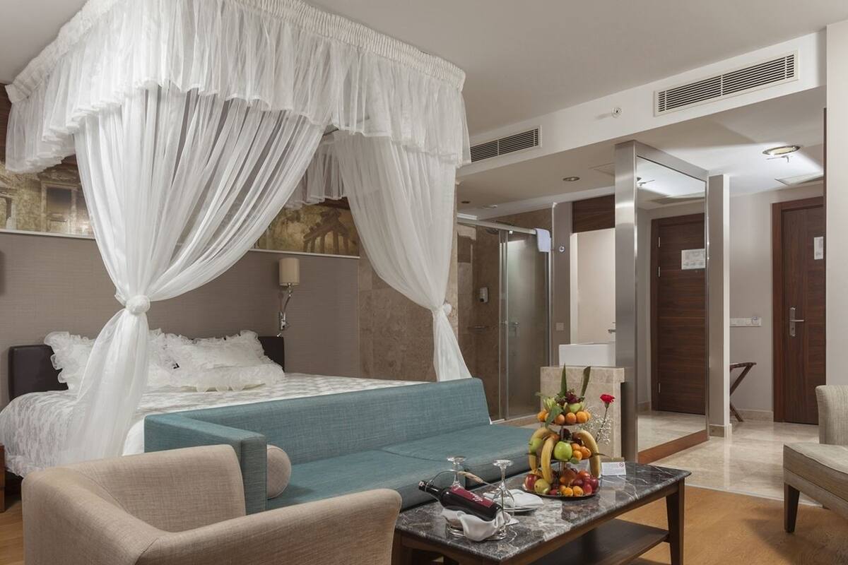 A bedroom with a four-poster bed, a sofa, a coffee table with fruit and a bottle, and a bathroom visible in the background.