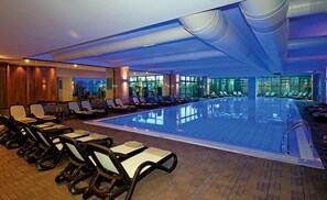 A large indoor swimming pool with lounge chairs and a sunbed area, surrounded by large windows and a modern ceiling design. - Commodore Elite Suites & Spa - All Inclusive - Adult Only (Manavgat)