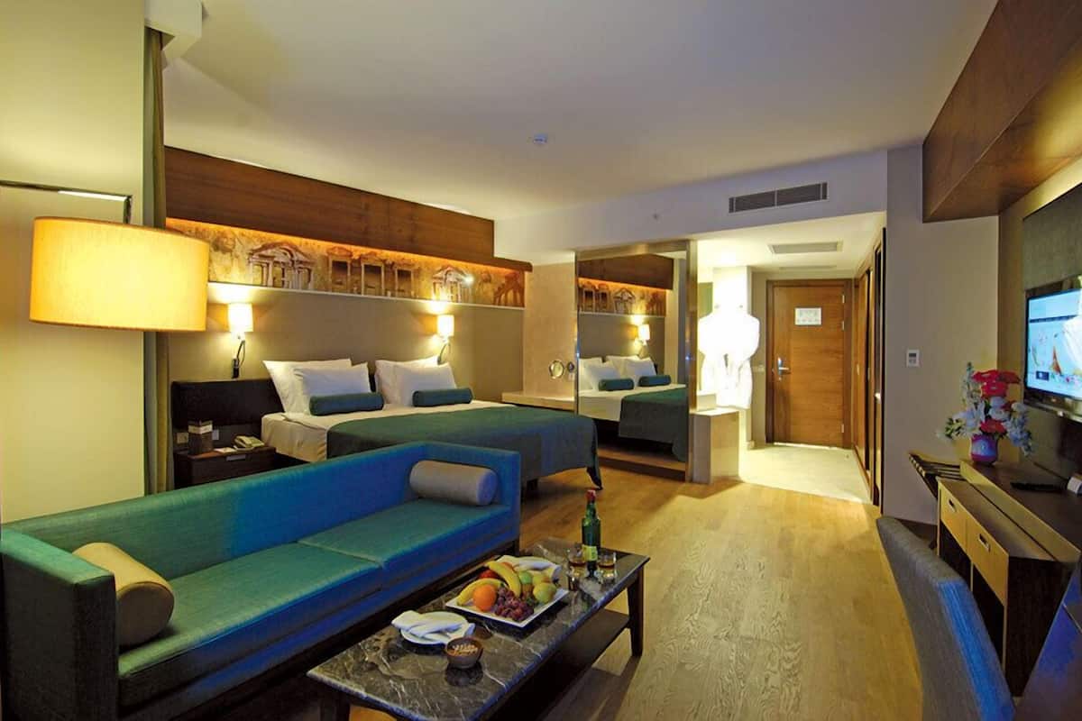 A hotel room with a large sofa, a smaller sofa, a desk with a TV, and a bed with two pillows.