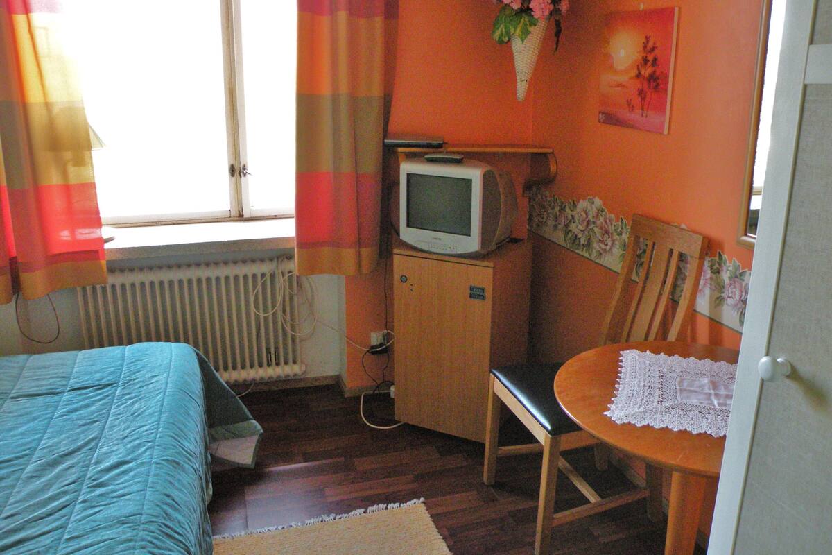 Une chambre à coucher avec un lit, un téléviseur posé sur un meuble en bois, une chaise et une petite table recouverte d’une nappe en dentelle.