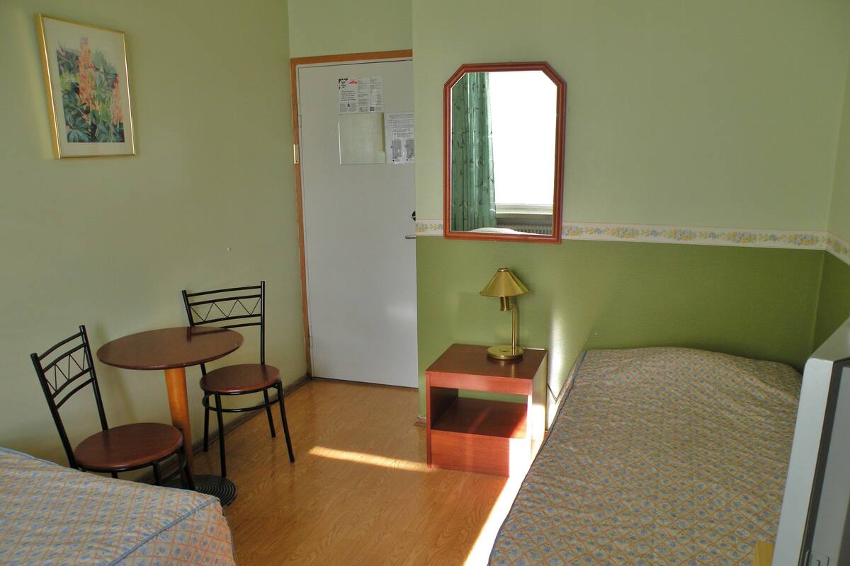 Une petite pièce simple, avec deux lits, une table et des chaises. Il y a une fenêtre et un miroir est accroché au-dessus de la table.