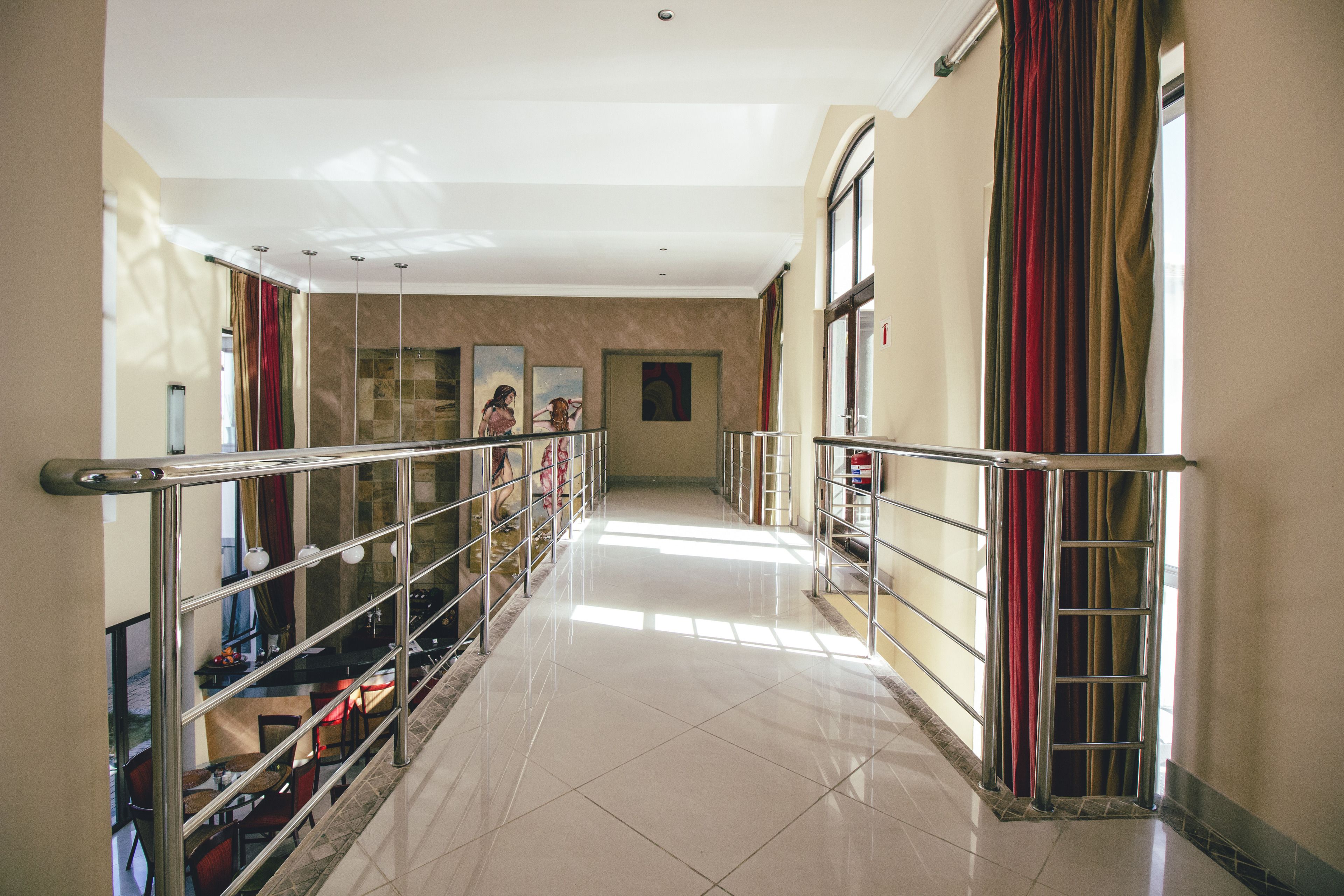 Corredor de hotel com guarda-corpo de vidro, piso ladrilhado e grandes janelas com cortinas.