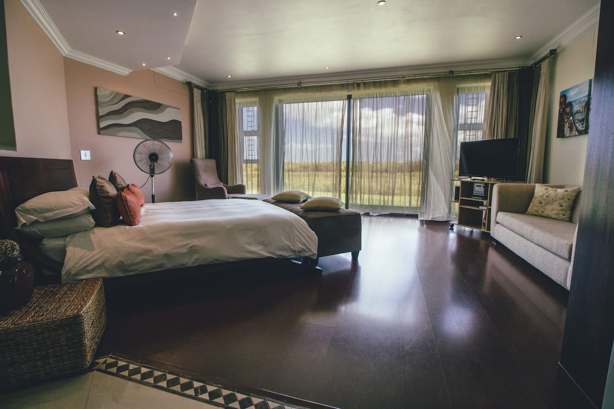 A spacious hotel room with a large bed, a sofa, a TV, and a view of the outdoors through the windows.