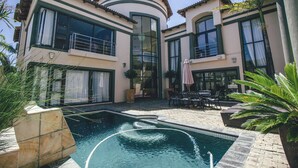 Una casa de dos pisos con piscina, mobiliario de exterior y plantas en macetas.