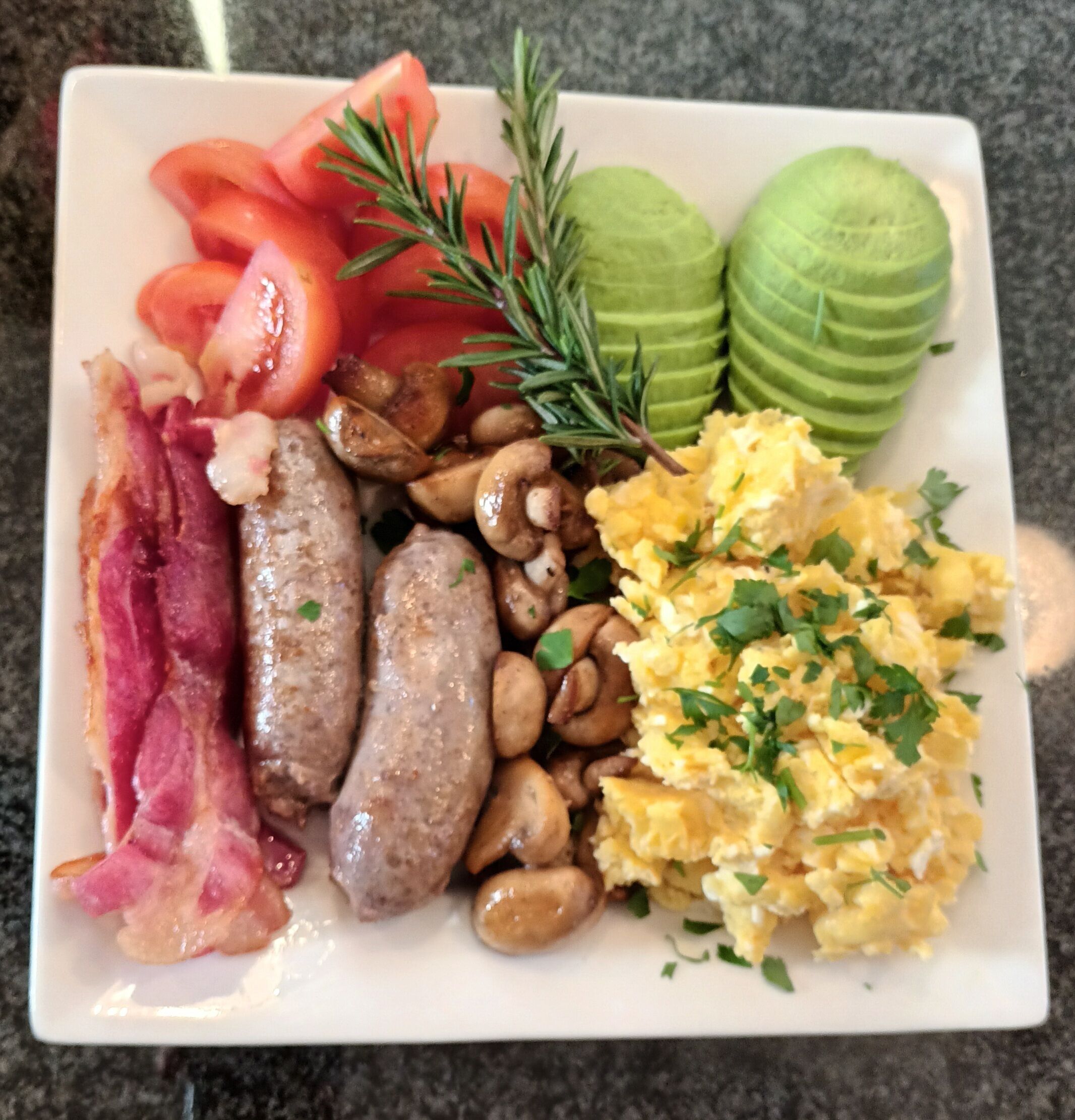 A plate with bacon, sausages, mushrooms, scrambled eggs, tomatoes, and avocado slices.