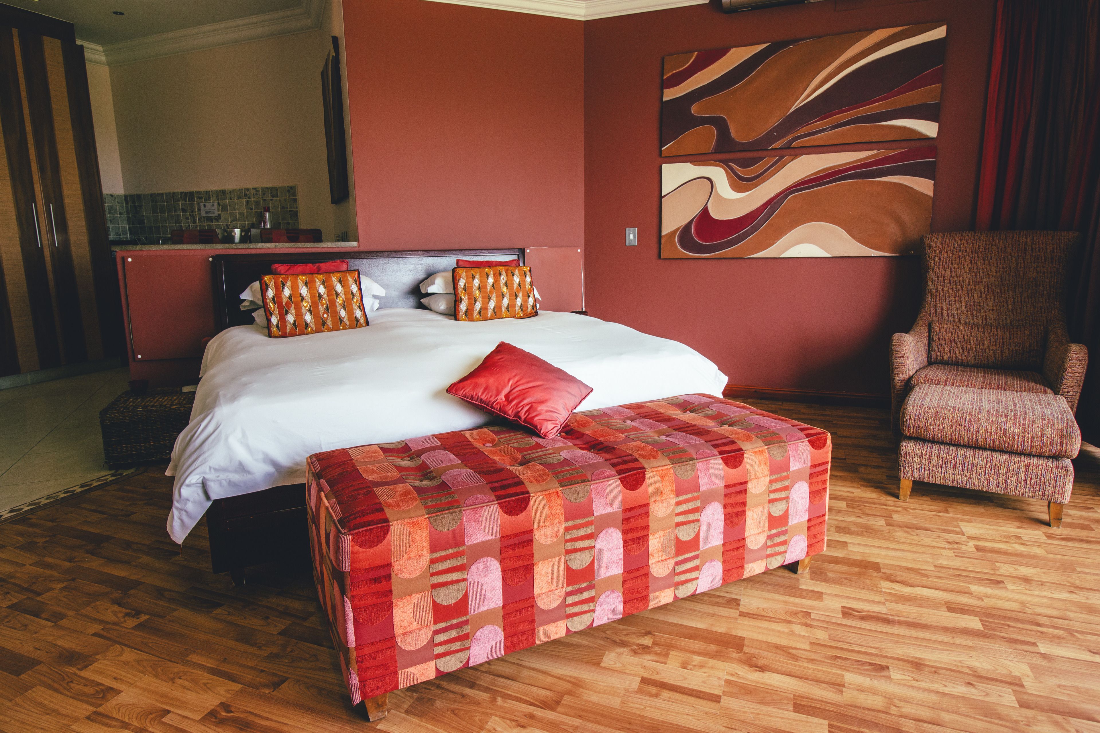 A hotel room with a large bed, a red and white patterned ottoman, a red armchair, and a kitchen area in the background.
