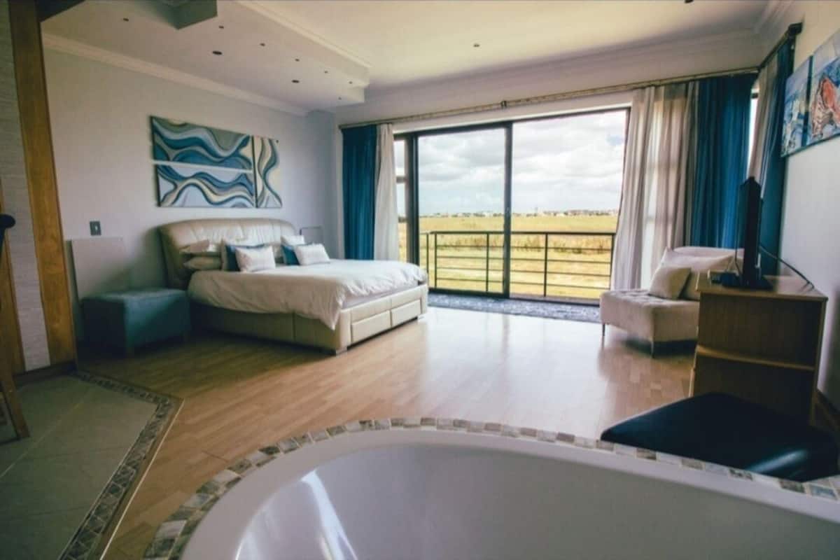 A spacious bedroom with a large bed, a sitting area, a bathtub, and a balcony with a view of the countryside.