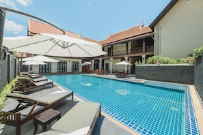 Piscina com espreguiçadeiras e guarda-sóis, cercada por um edifício com telhado em azulejos.