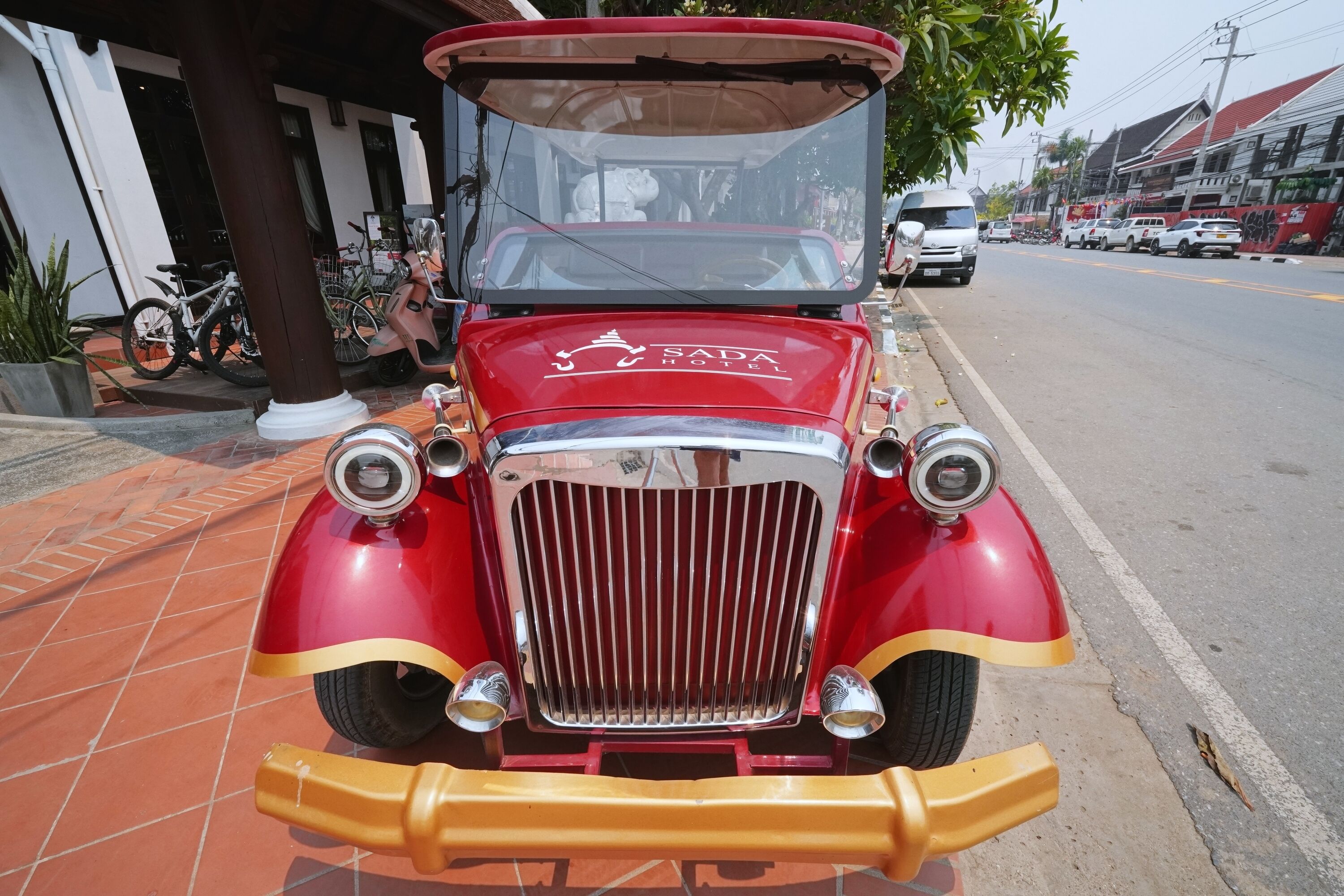 Een klassieke rode auto geparkeerd voor een gebouw met een bord waarop staat 'Sada Hotel'.