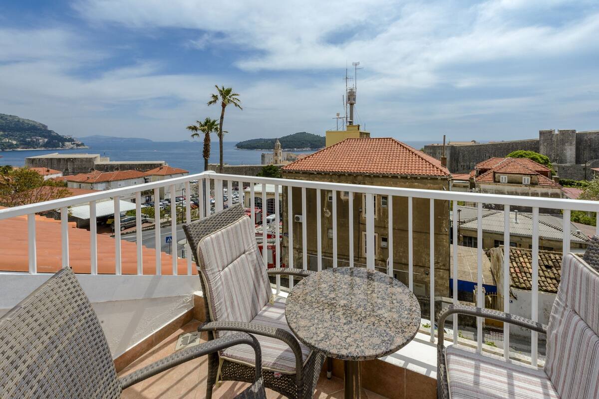 A balcony with wicker furniture and a round table, overlooking a coastal town with historic buildings and a clear view of the sea.