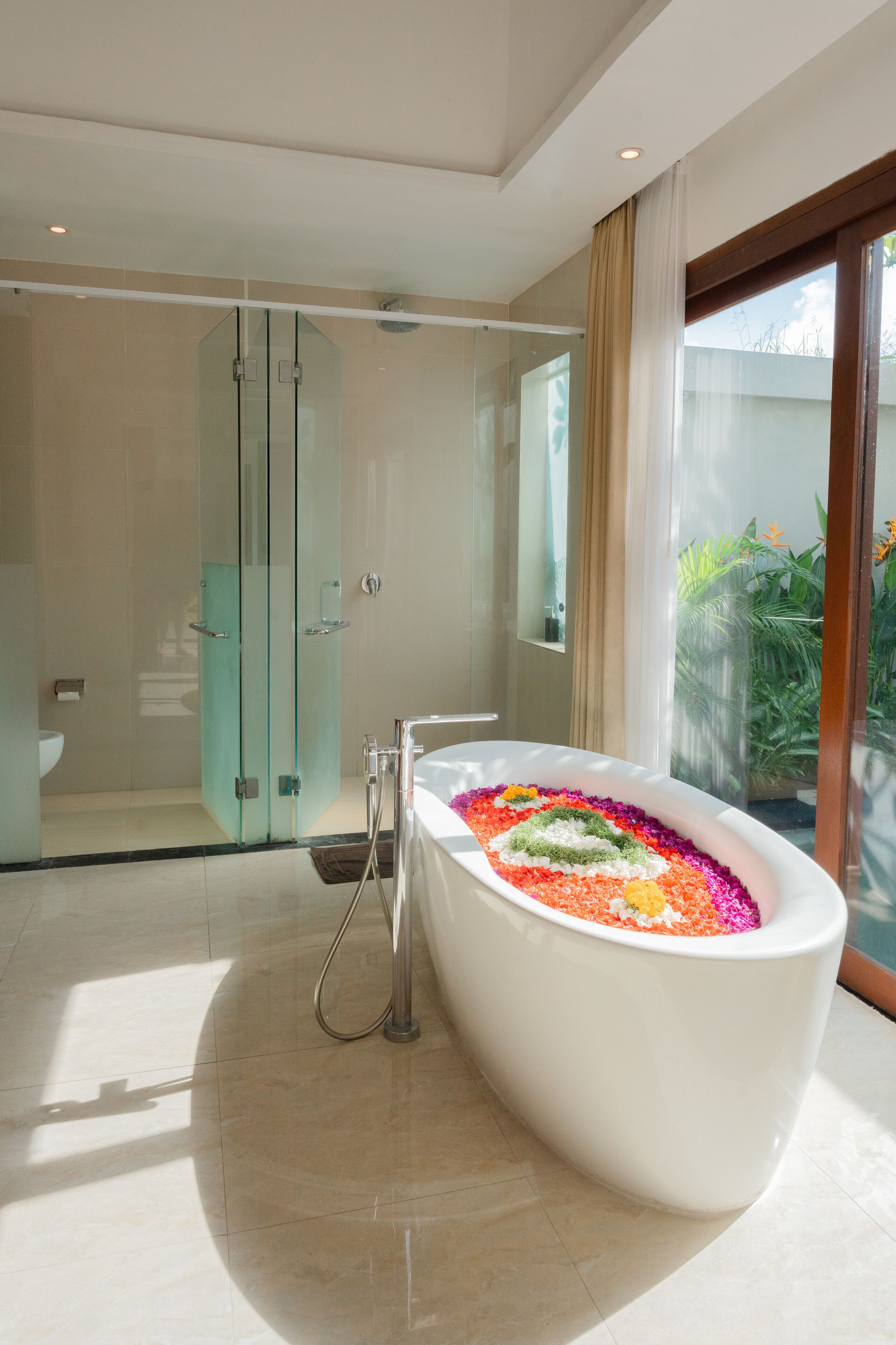 A modern bathroom with a freestanding bathtub filled with colorful bath salts, a glass-enclosed shower, and a large window providing natural light.