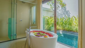 Een moderne badkamer met een vrijstaand bad gevuld met kleurrijke bloemen, een douchecabine van glas en uitzicht op een zwembad en groen.