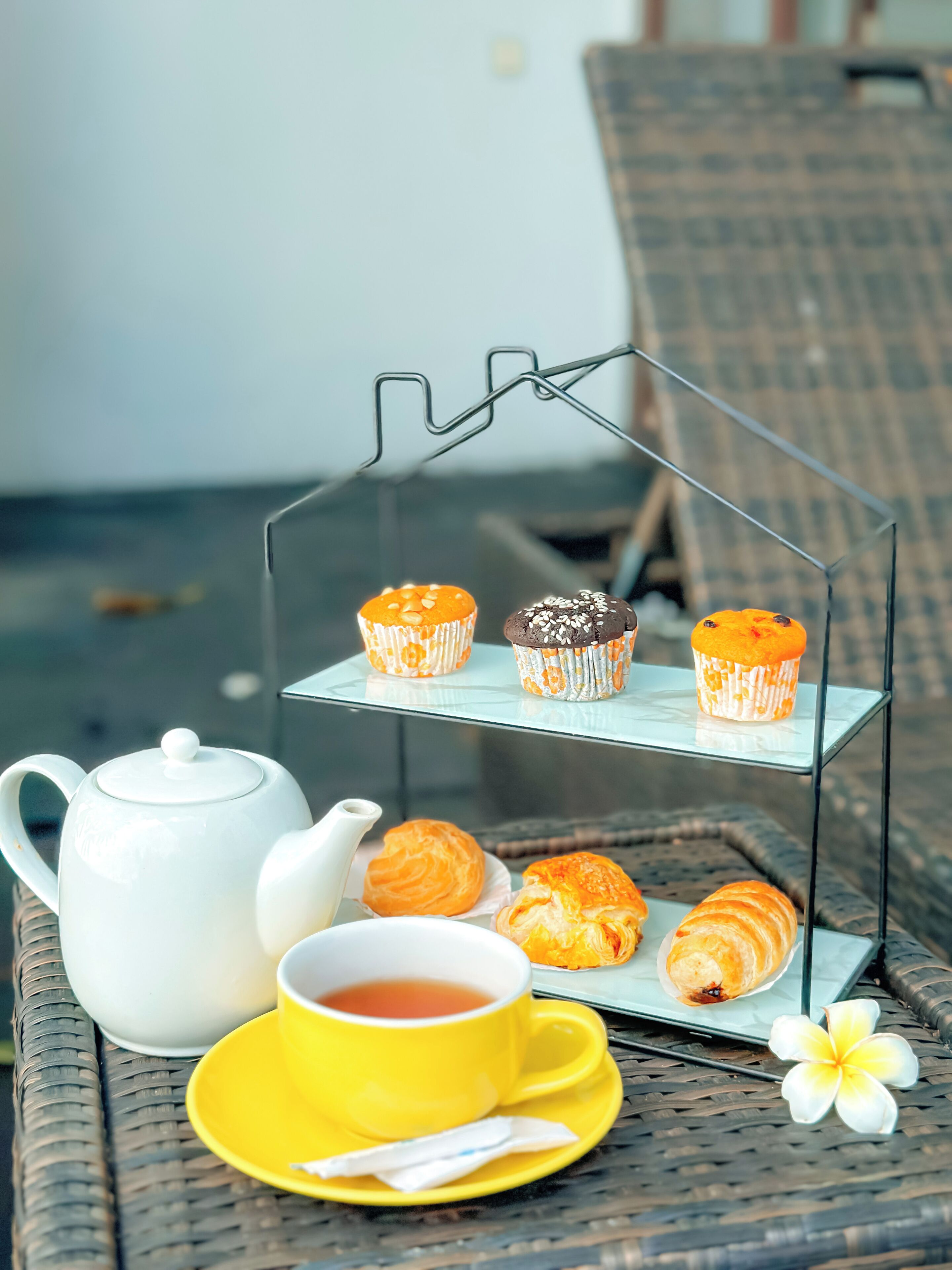 Een theeset met theepot en kopje op een rieten tafel, met gebakjes op een draadstandaard en een gele schotel.