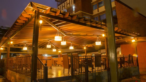 Un espacio exterior cubierto con estructura de madera, varias luces colgantes y un edificio de ladrillo al fondo.