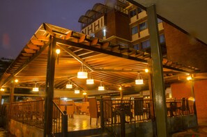 Un espacio exterior cubierto con estructura de madera, varias luces colgantes y un edificio de ladrillo al fondo.