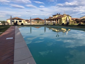 A swimming pool with a clear reflection, surrounded by buildings with red roofs and outdoor seating areas. - Agriturismo Il Boschetto (Ornago)