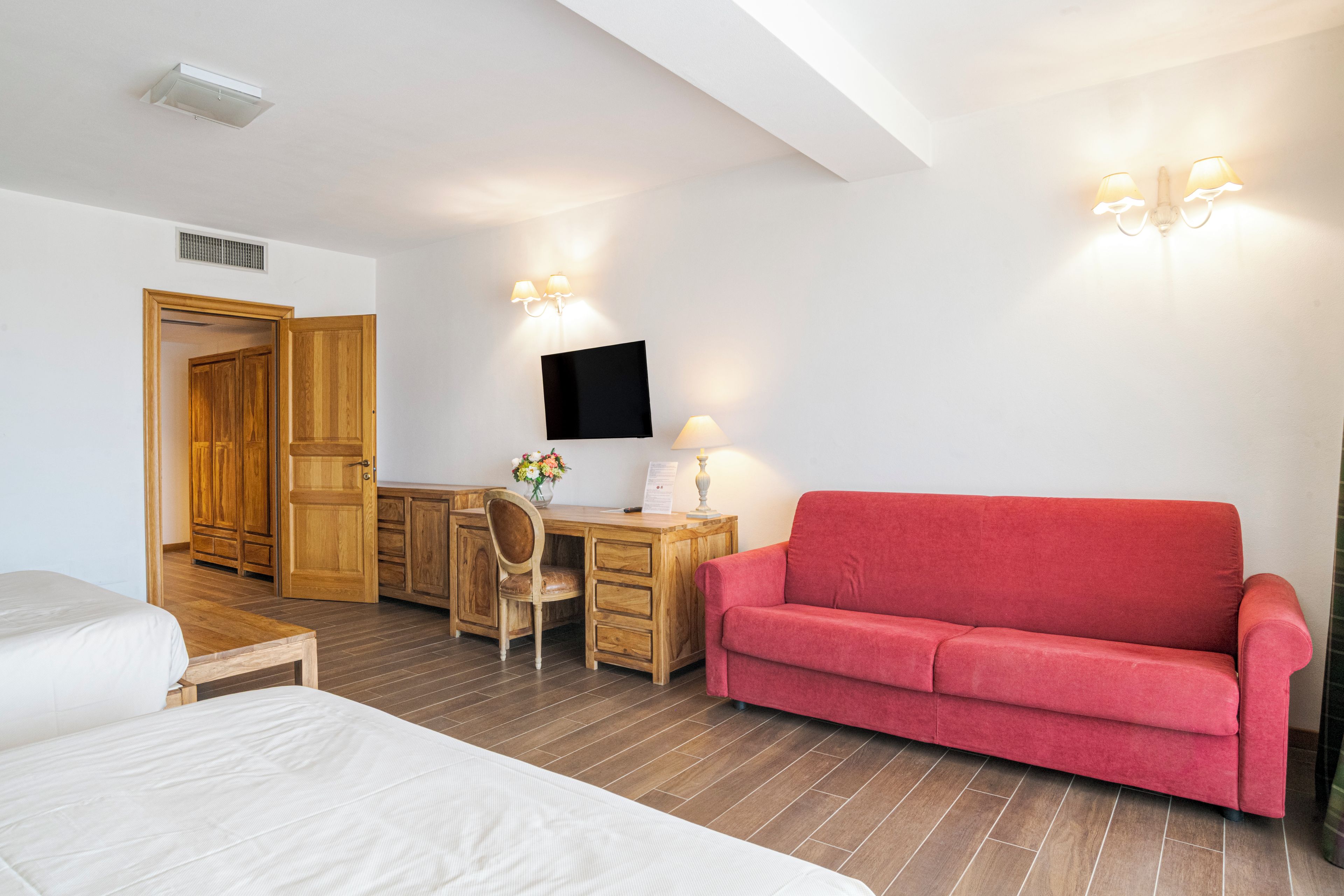 Une chambre d’hôtel avec un bureau en bois, un canapé rouge, un lit et un téléviseur fixé au mur.