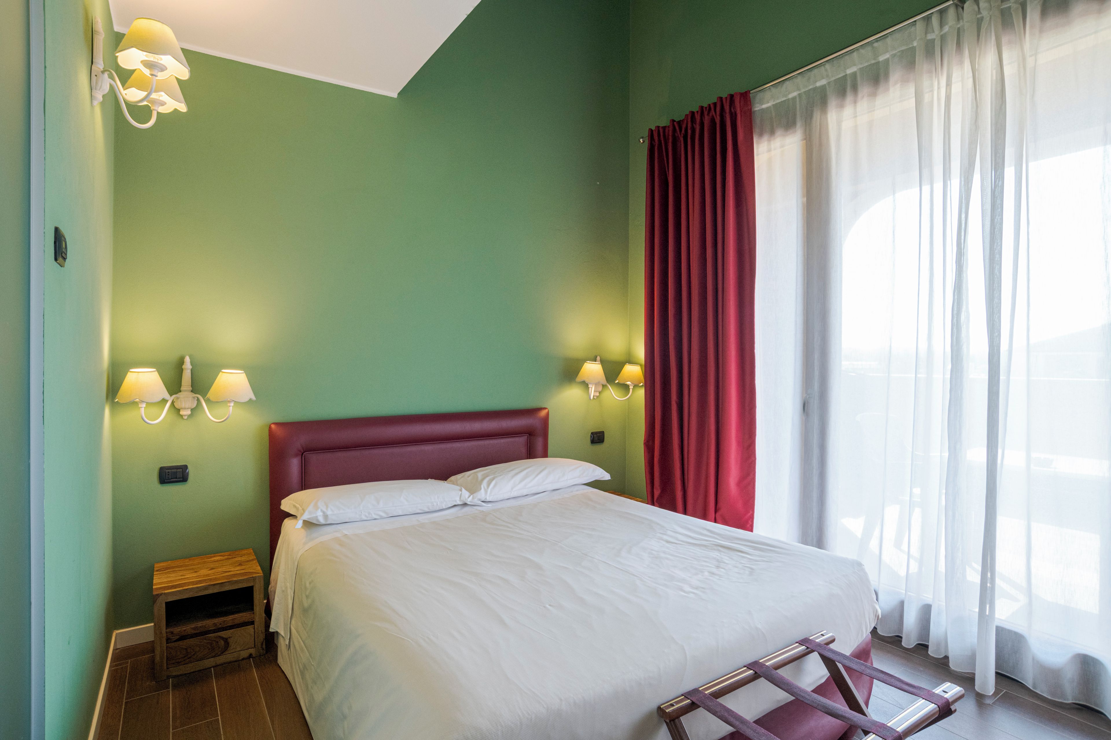 A hotel room with a bed, two bedside lamps, a nightstand, a wooden bench, and a window with curtains.