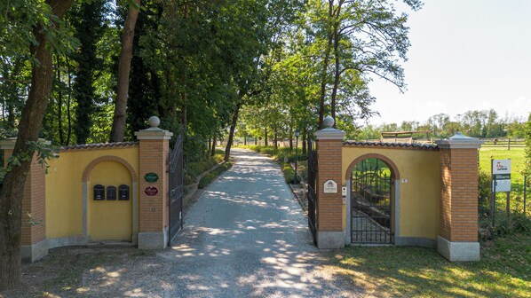 A paved pathway leading through a gated entrance with a brick wall and arched gate, surrounded by trees. - Agriturismo Il Boschetto (Ornago)
