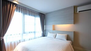 A hotel room with a large bed, a headboard with built-in lighting, a window with curtains, and an air conditioning unit on the wall. - The Present Sathorn (Bangkok)