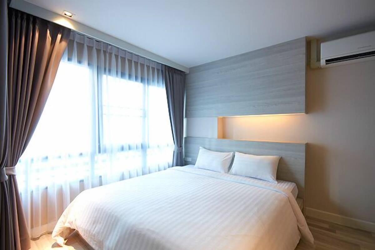 Ein Hotelzimmer mit einem großen Bett, einem Kopfteil mit integrierter Beleuchtung, einem Fenster mit Vorhängen und einer Klimaanlage an der Wand.