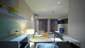 A modern hotel room with a kitchenette, a sink, a chair, a small table, a TV, and a window with curtains. - The Present Sathorn (Bangkok)