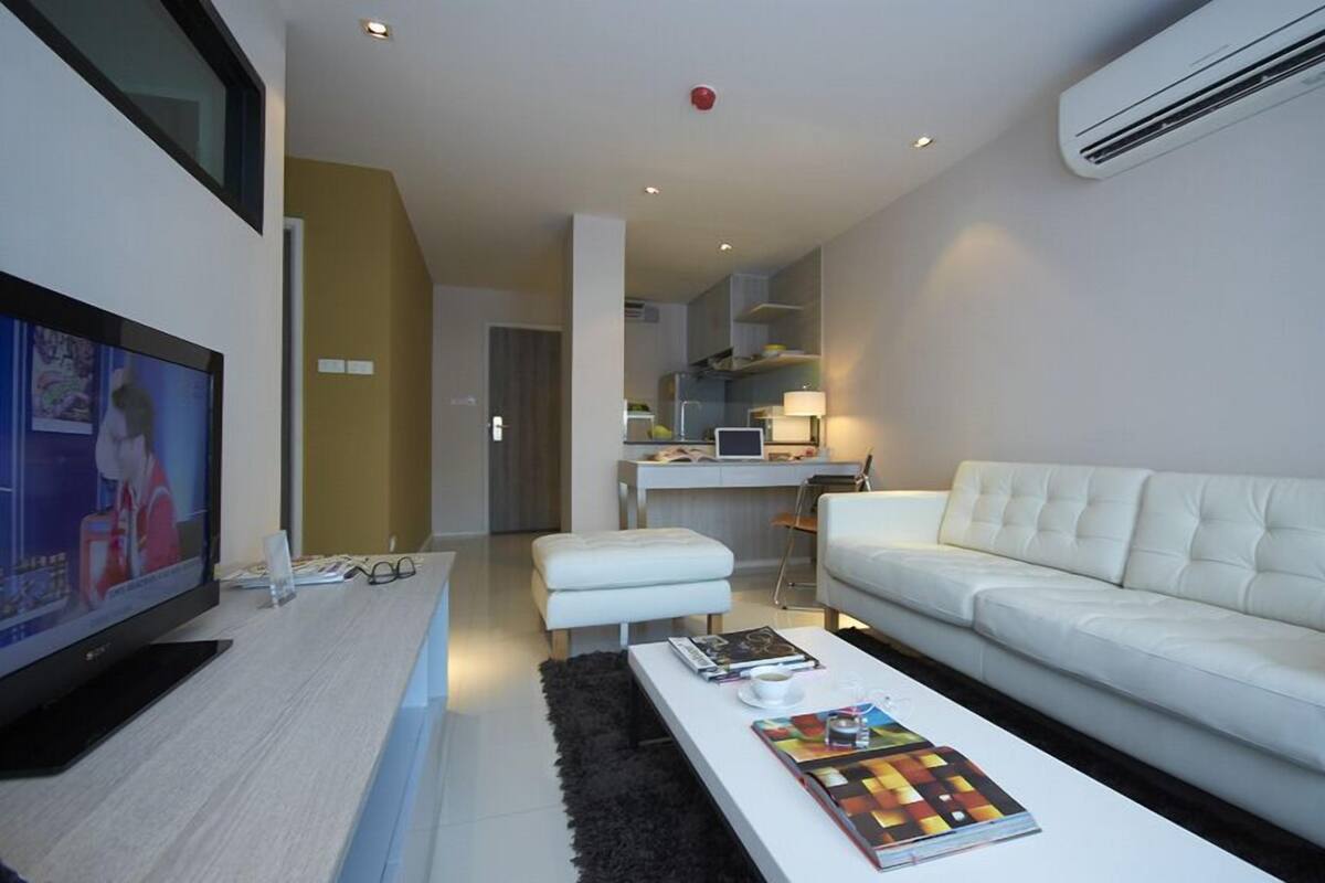 Ein modernes Wohnzimmer mit Flachbildfernseher, einem weißen Sofa, einem Couchtisch mit Büchern und einer Küche im Hintergrund.