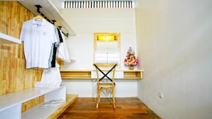 A hotel room with a wooden floor, a white wall, a wooden shelf with clothes, a mirror, and a small flower arrangement.