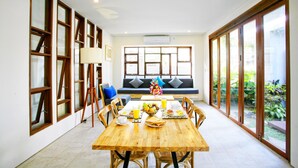 A dining area with a wooden table and chairs, a sofa, and large windows with a view of greenery.