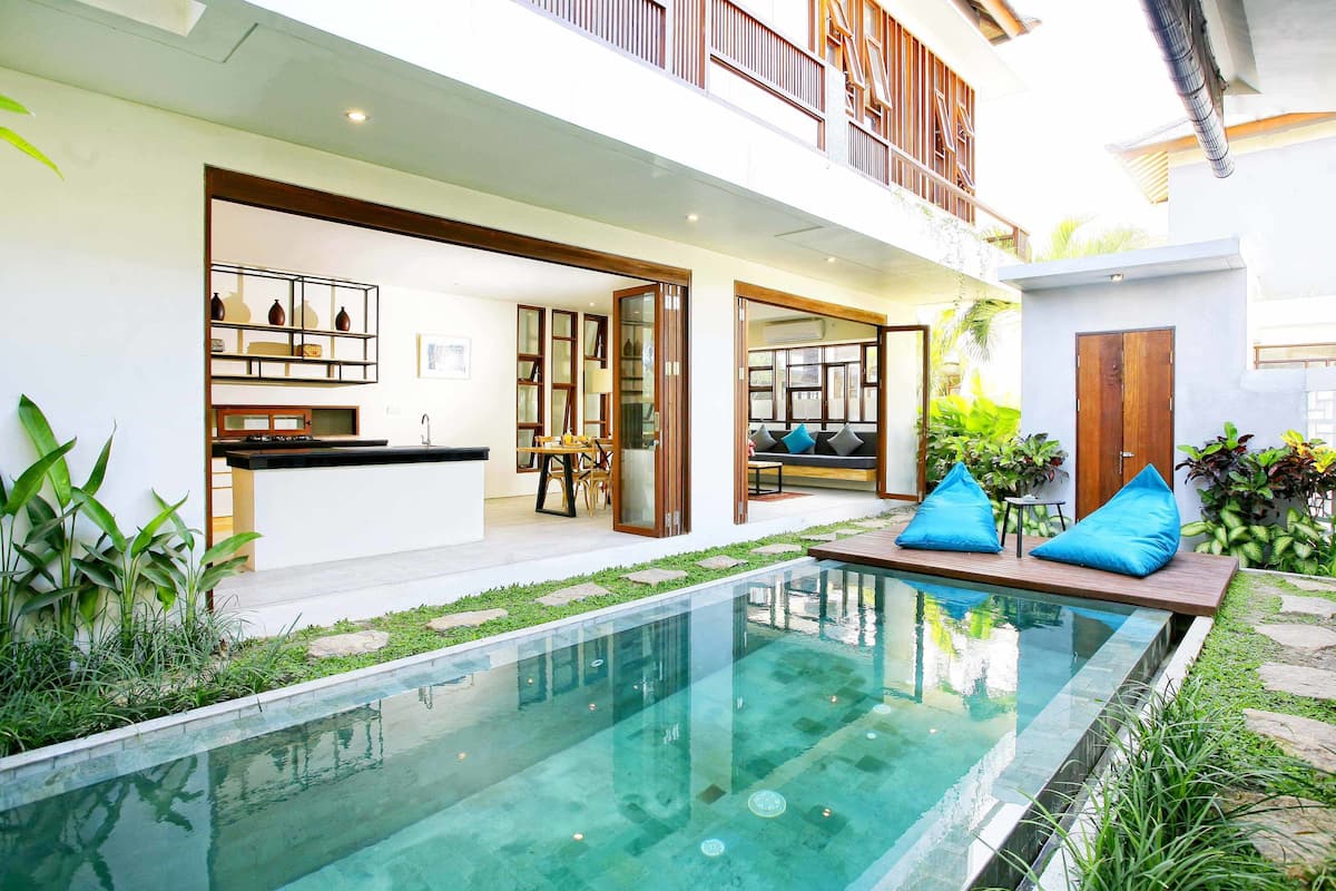 Uma casa moderna com piscina, varanda e cozinha visível por uma porta aberta.