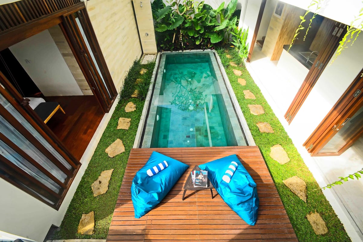 Uma área moderna de piscina ao ar livre, com um deck de madeira, duas áreas de estar azuis com almofadas e uma pequena mesa com uma revista.