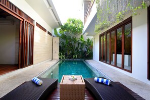 A poolside area with two wicker lounge chairs, a small table with a flower, and a glass-enclosed pool.