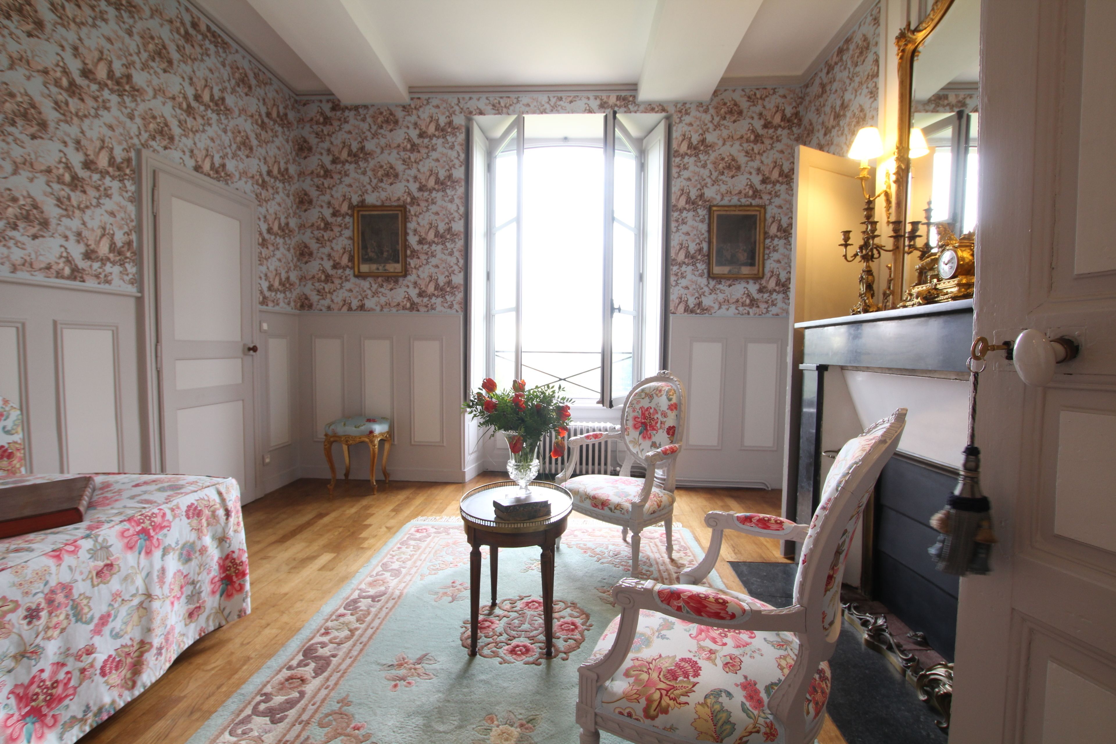 Una stanza con carta da parati floreale, pavimento in legno, un tavolo rotondo con un vaso, due sedie, un letto con lenzuola a motivi floreali e un camino con specchio e candelabro.
