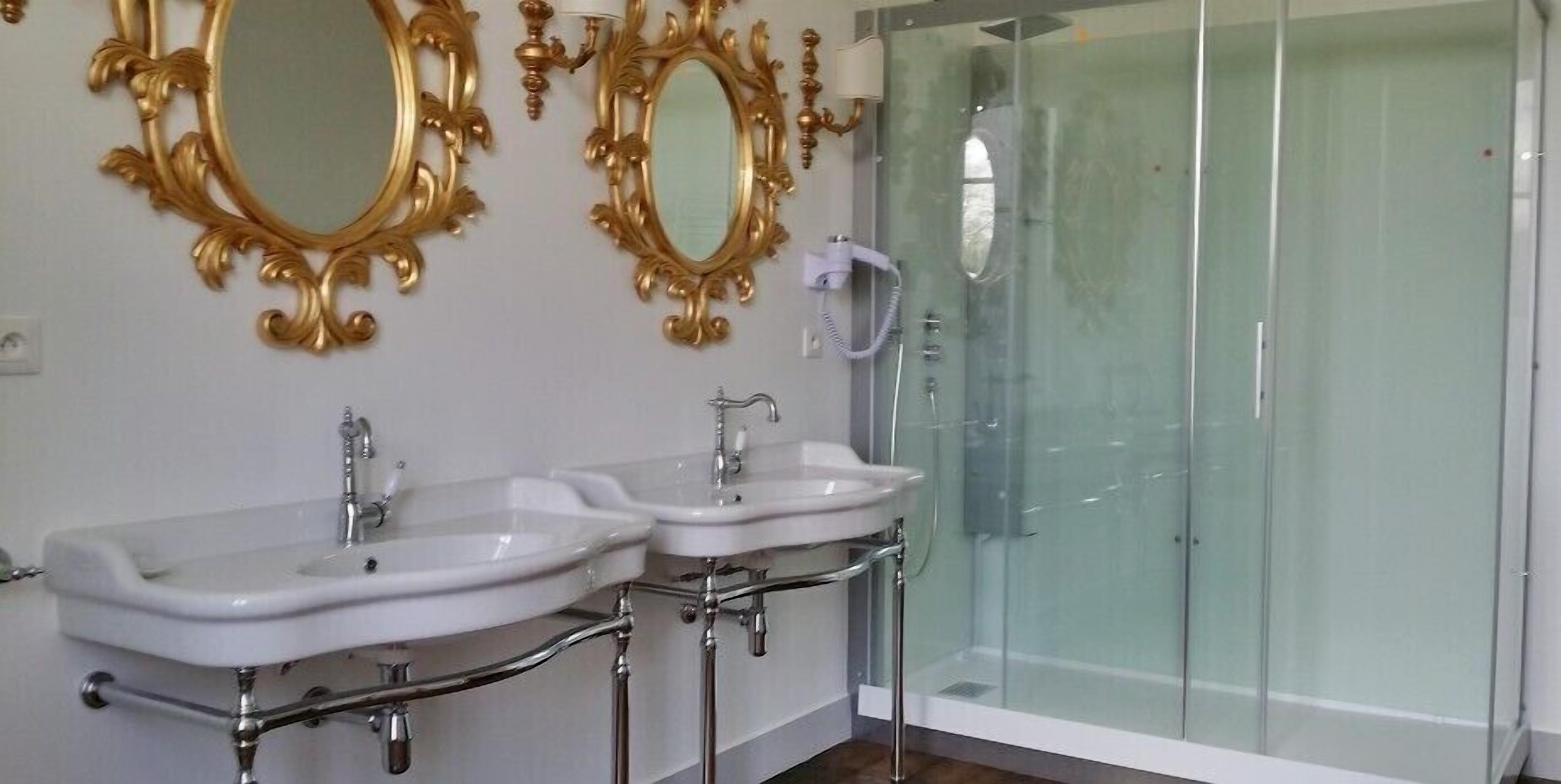 Un baño con dos lavamanos, una cabina de ducha de vidrio y espejos con marcos dorados ornamentados.