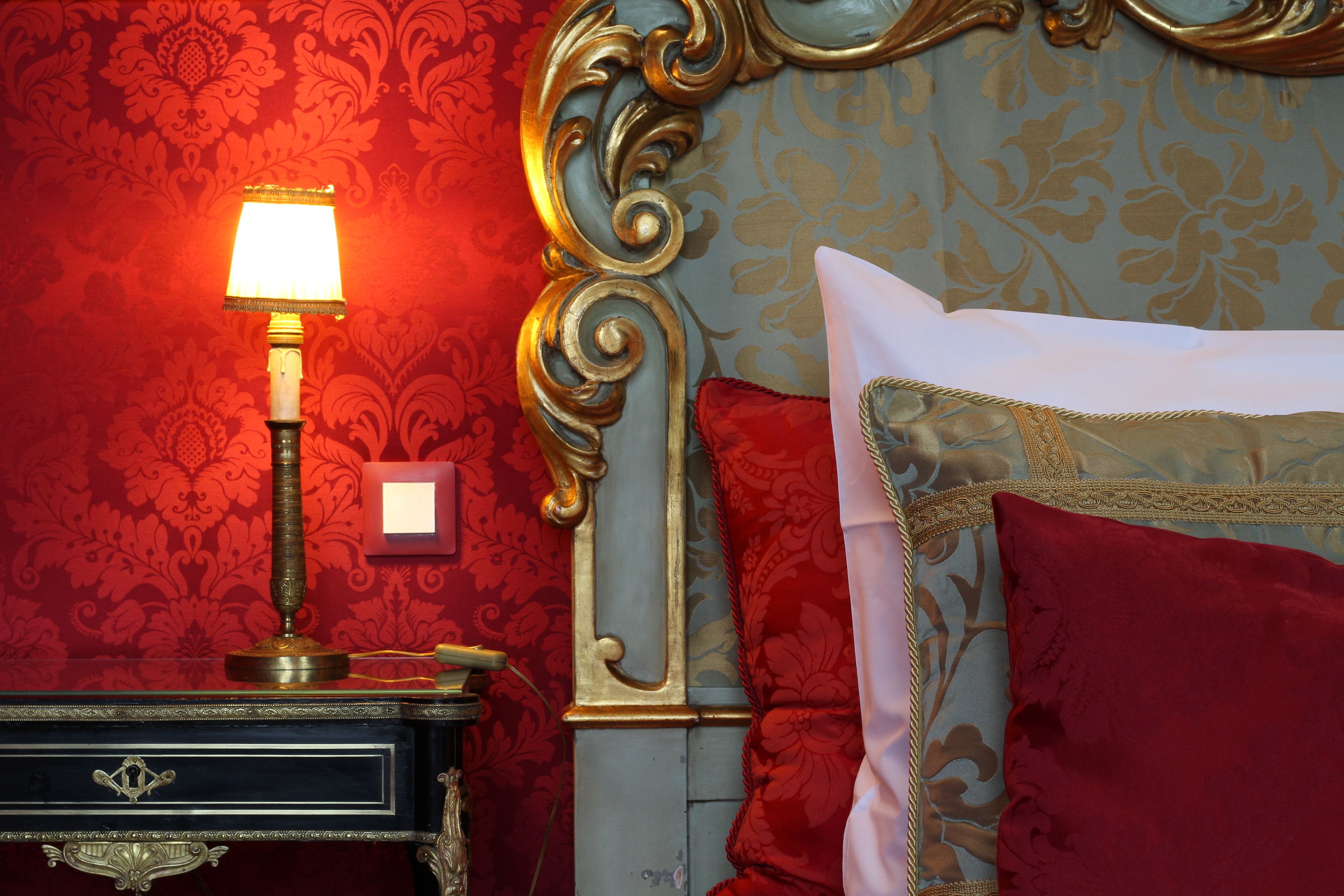 A bed with red and gold bedding, a nightstand with a lamp, and ornate wall decor.