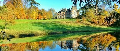 A large, historic house with a pond in front, surrounded by autumn-colored trees.