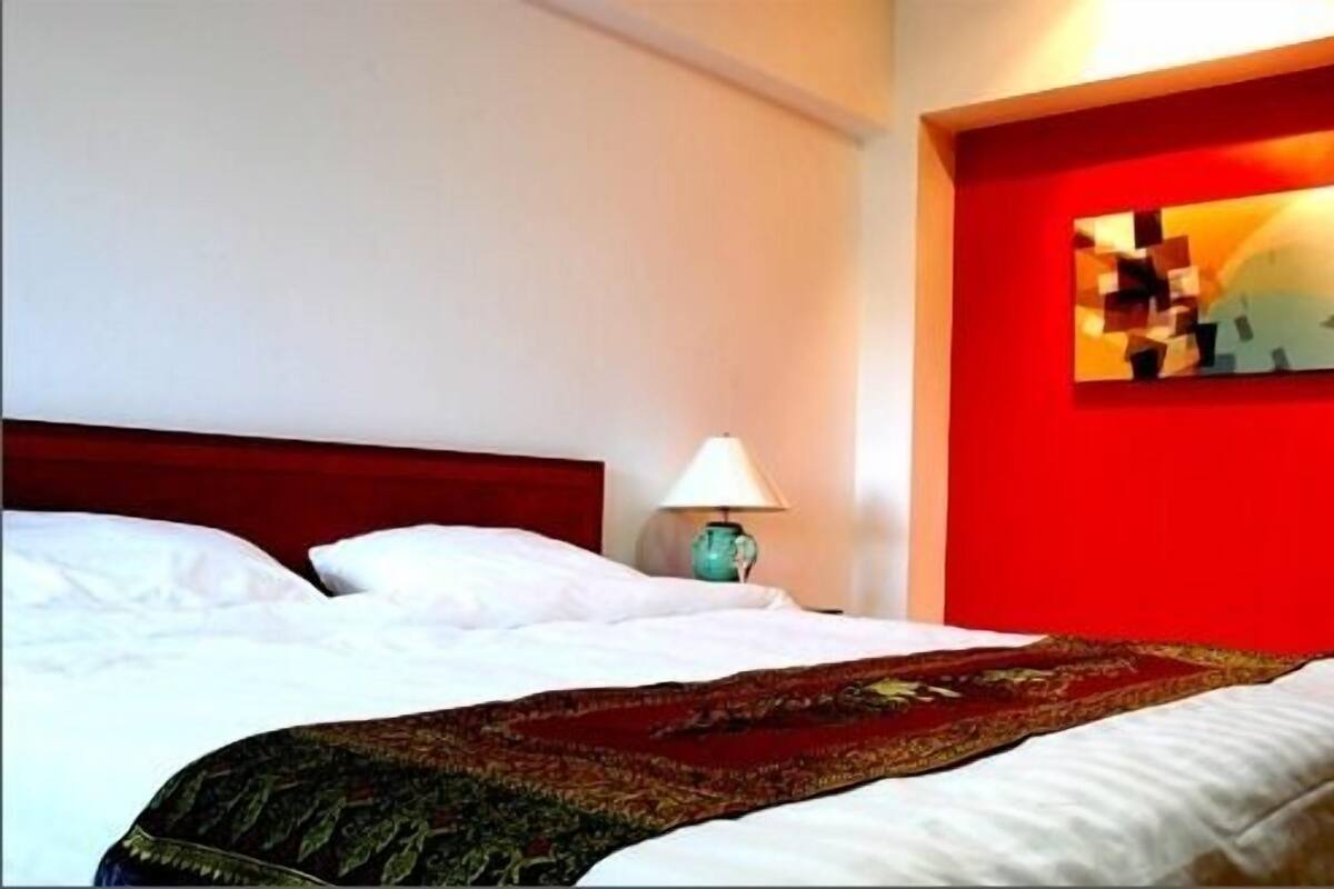 Una habitación de hotel con cama, mesita de noche con lámpara y pared de acento roja con un cuadro.