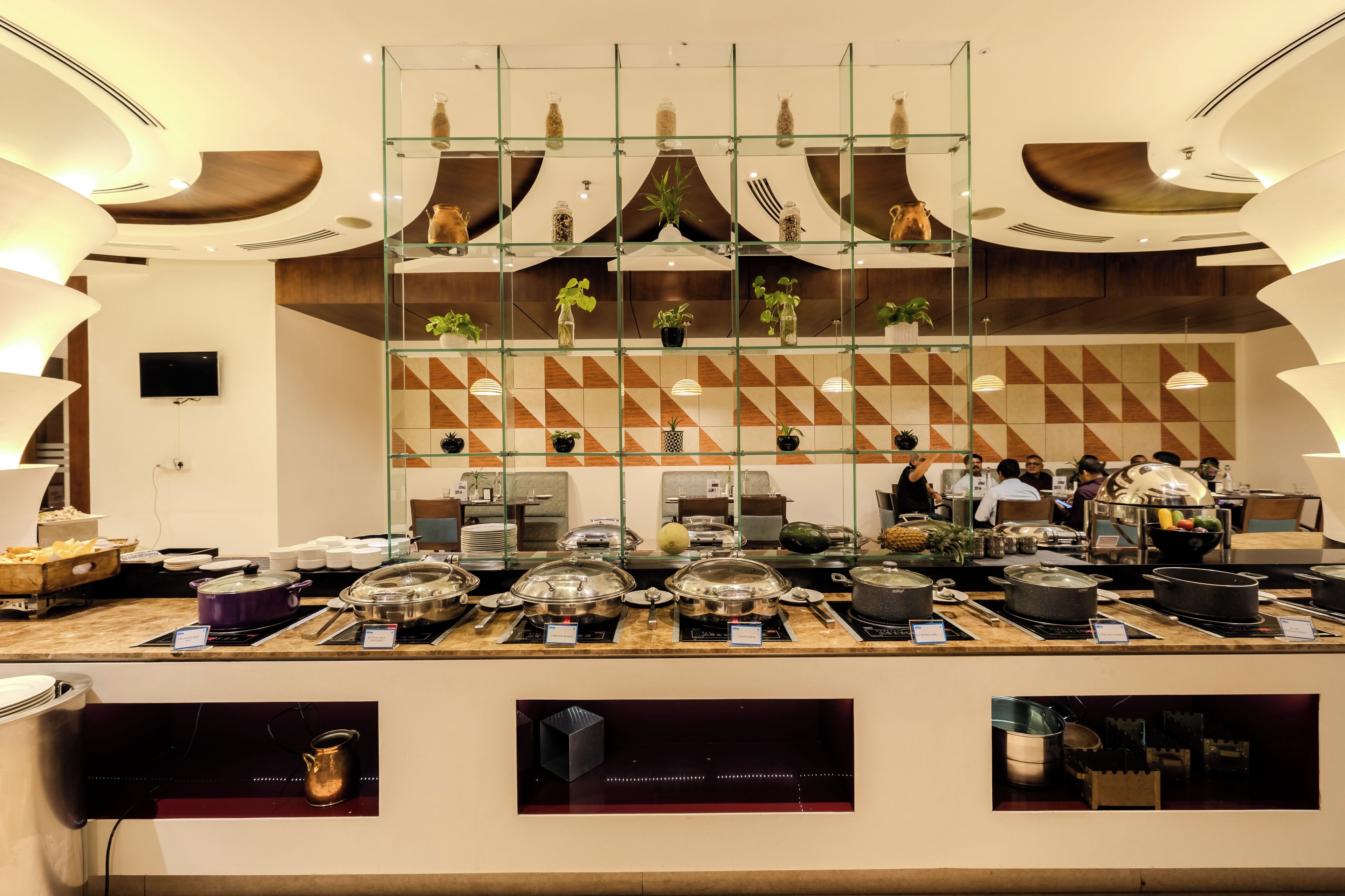 En buffetafdeling med flere serveringsstationer, glasskabe med dekorative genstande og en væg med geometrisk mønster.