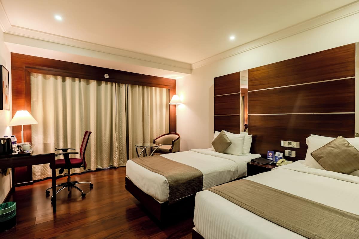 Habitación de hotel con dos camas, un escritorio con silla, una lámpara y una ventana con cortinas.