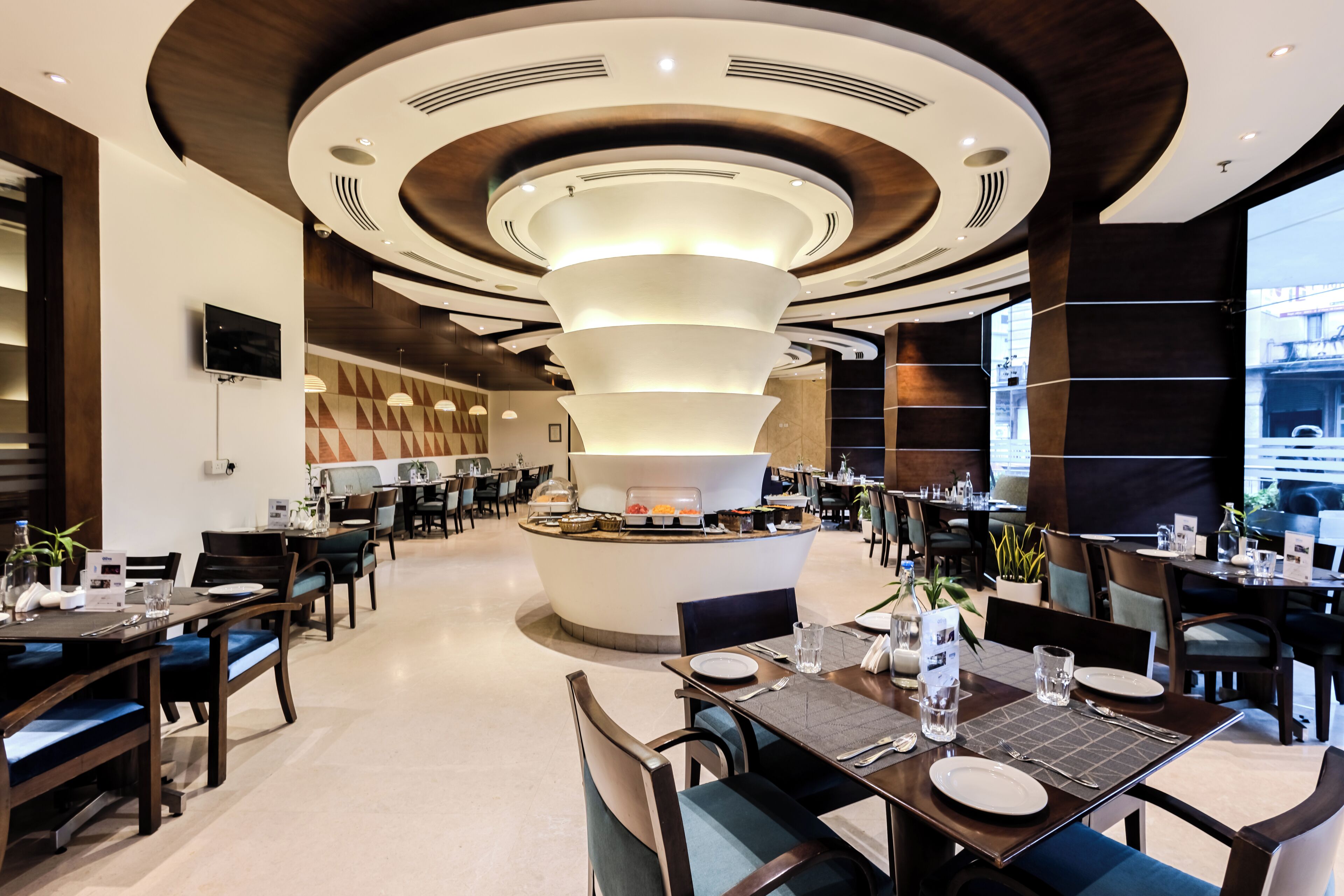 Un moderno vestíbulo de hotel con un mostrador de recepción central, zona de comedor con mesas y sillas, y un amplio ventanal que ofrece vistas al exterior.