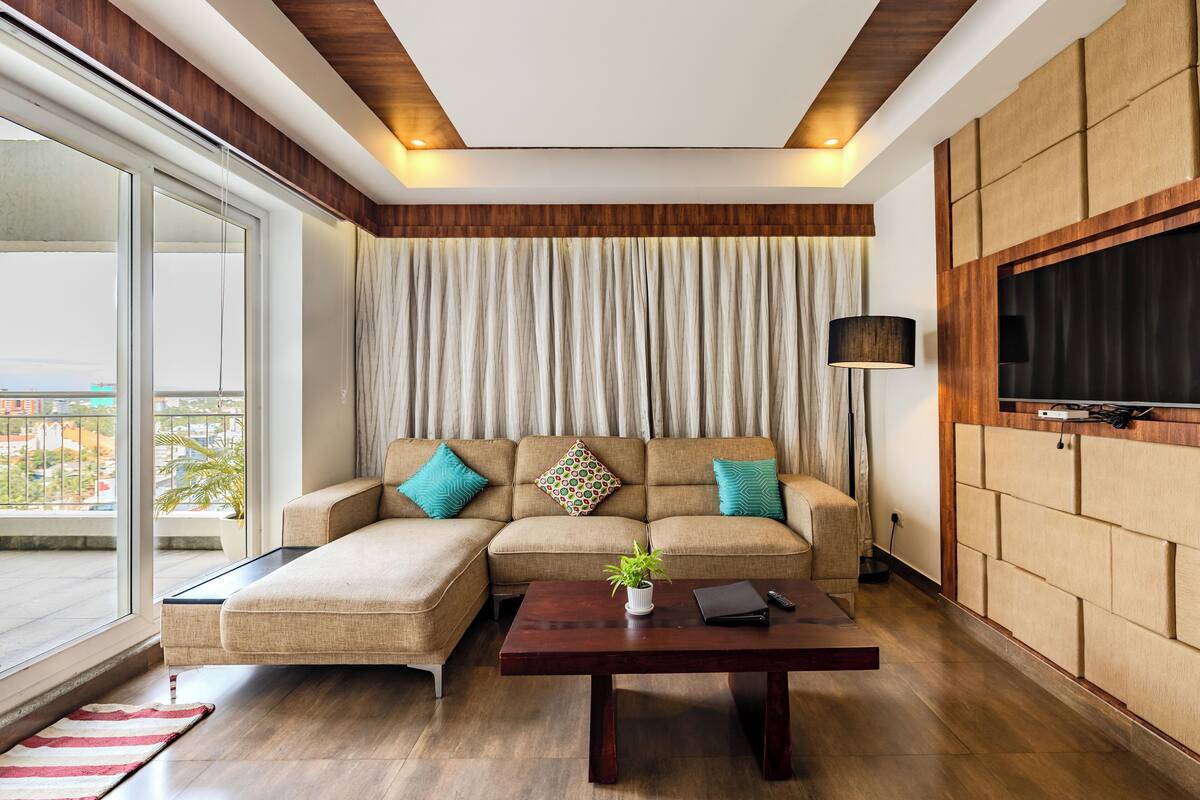 Una sala moderna con un sofá seccional, una mesa de centro, un televisor y un amplio ventanal con cortinas.
