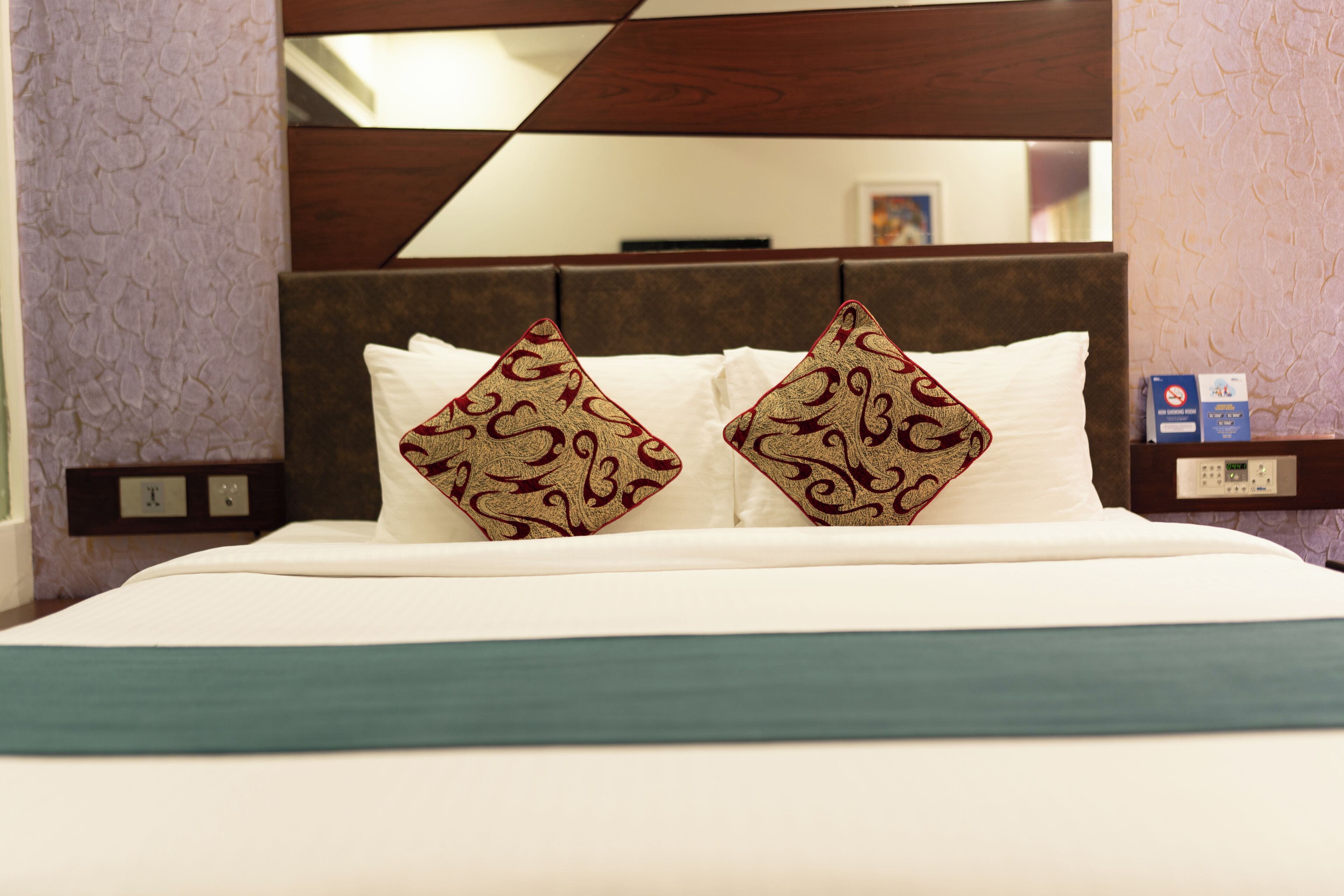 A hotel room with a bed, two decorative pillows, a nightstand with a clock, and a wall-mounted control panel.