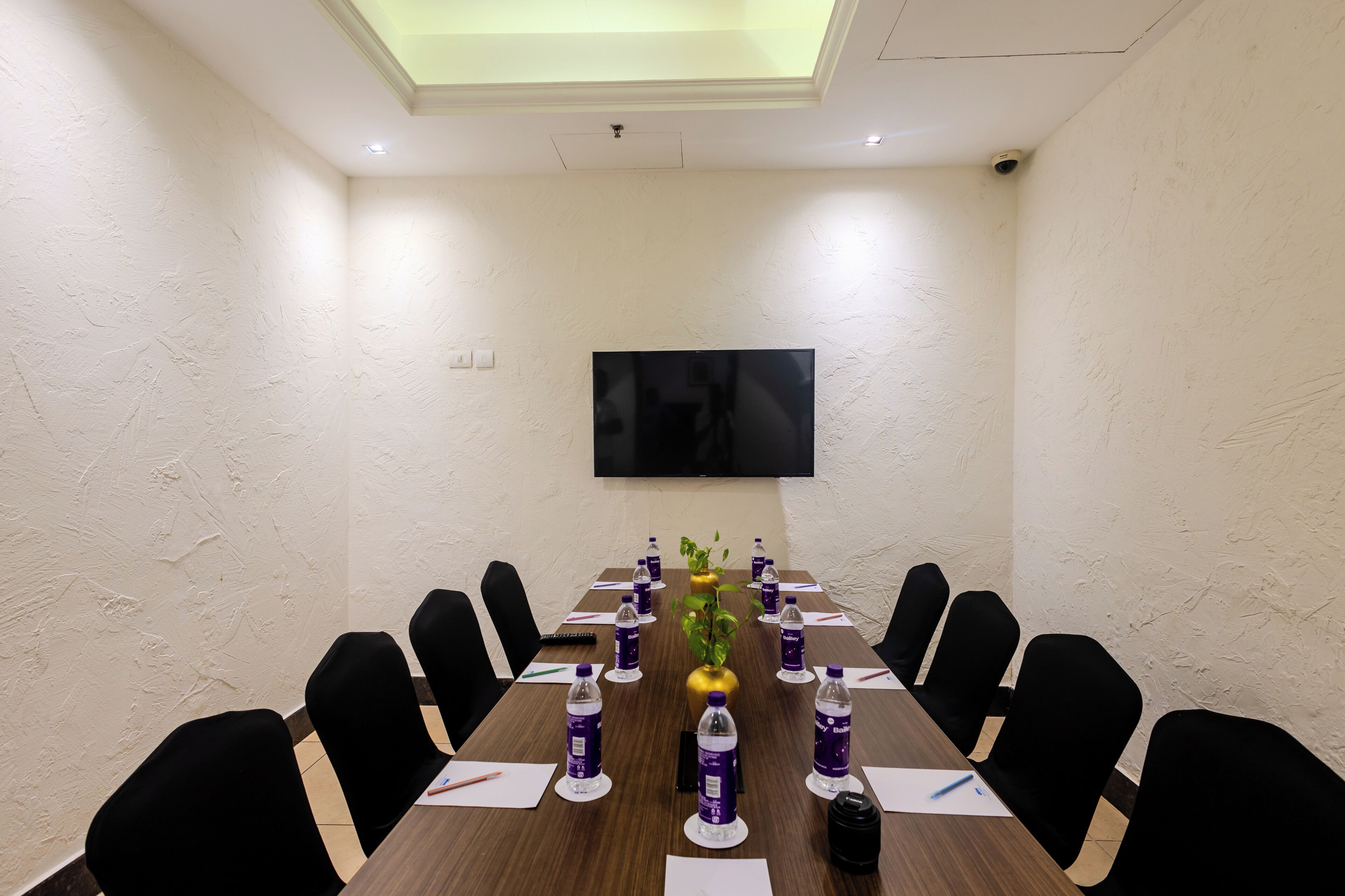 Un salón de conferencias con una larga mesa, sillas negras, un televisor de pantalla plana y botellas de agua sobre la mesa.