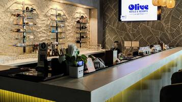 Un espace bar avec des étagères remplies de bouteilles et de verres, un écran numérique affichant le nom « Olive Hotels & Resorts », et un design moderne avec un mur texturé et des touches de couleur jaune.
