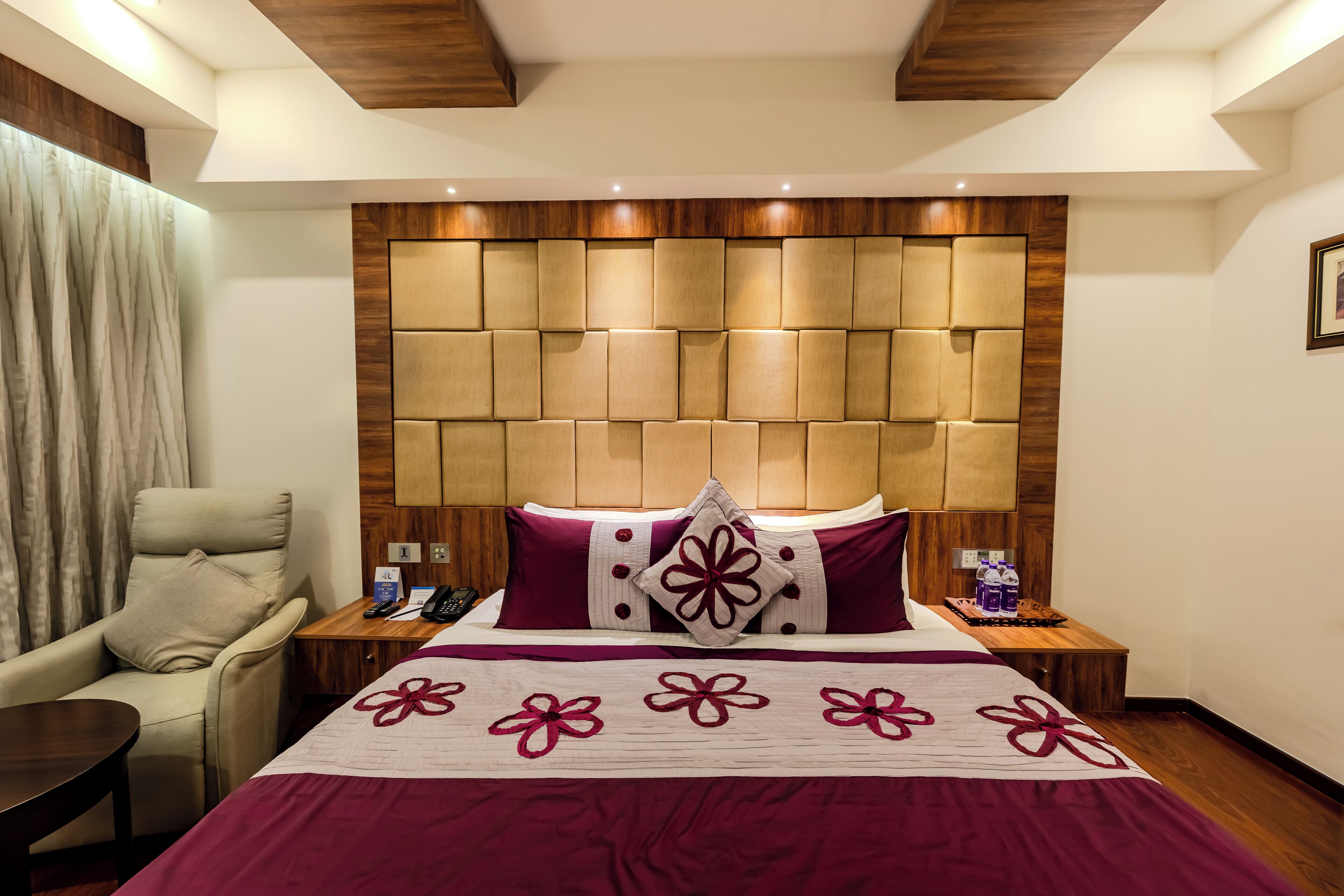 A hotel room with a large bed, a wooden headboard, a chair, a nightstand, and a bedside table.