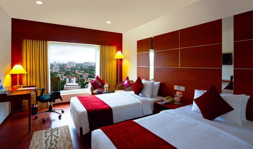 Habitación de hotel con dos camas, un escritorio y una silla. Cuenta con un ventanal con cortinas, y un diseño moderno con paneles de madera.