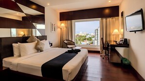 A hotel room with a large bed, a desk, a chair, a TV, and a view of the city through a large window. - Olive Downtown Kochi (Kanayannur)
