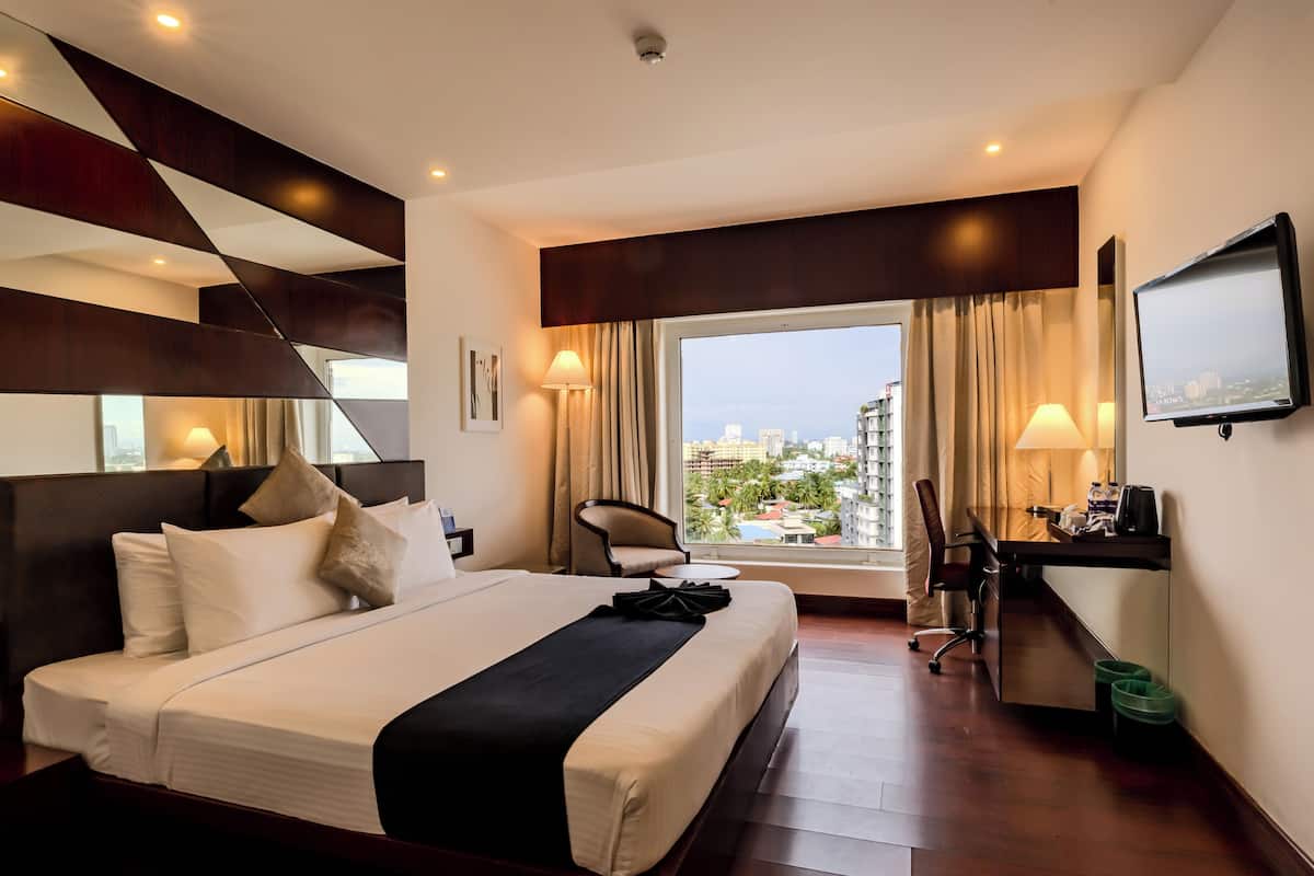 Habitación de hotel con una cama grande, un escritorio, una silla, un televisor y vistas a la ciudad a través de un ventanal.