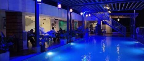 Una piscina interior con iluminación azul, una zona cubierta con asientos y una escalera que conduce a un nivel superior.